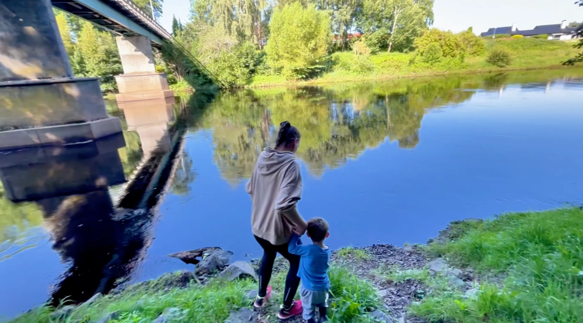 Bron vid Vallarnas Friluftsteater En vuxen och ett barn står vid en lugn flod med en bro i bakgrunden.