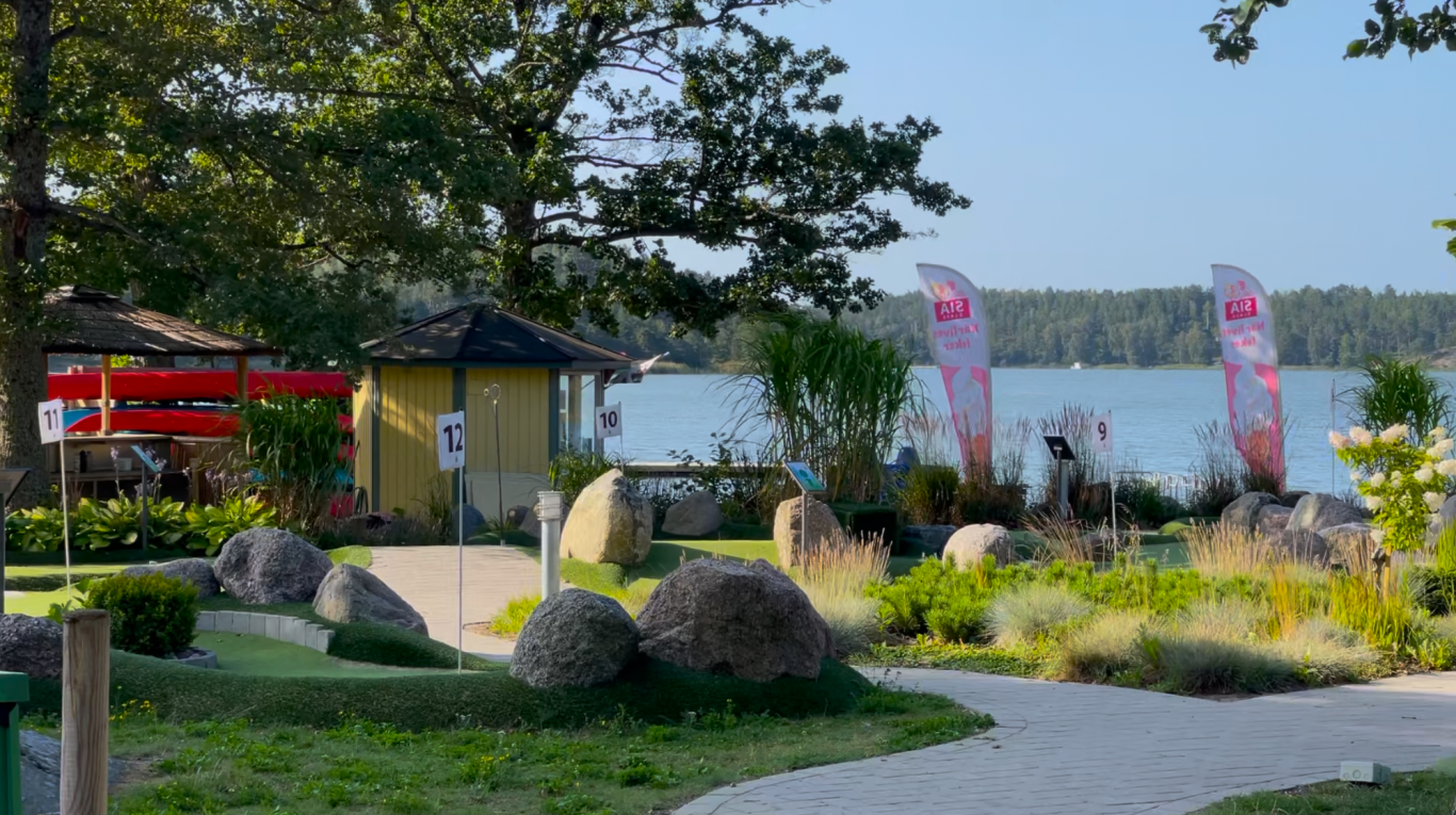 Minigolfbanan Landskap vid sjön med ett grönområde, stuga och två flaggor i bakgrunden.