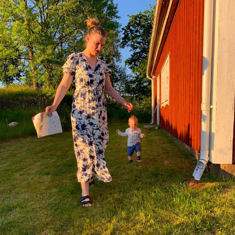 Kvinna i blommig klänning går på gräset med ett barn framför en röd stuga.