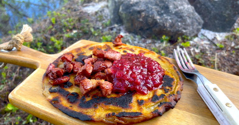 Kolbulle med stekt sidfläsk och lingonsylt på en träskärbräda. I bakgrunden en eldstad.