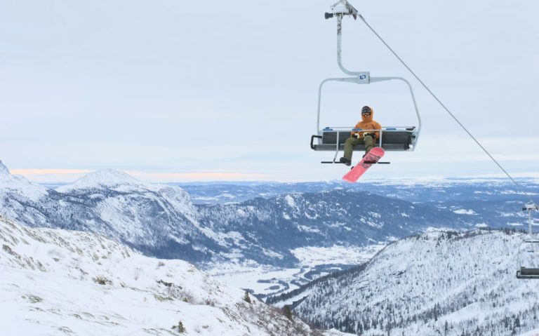 En snowboardåkare sitter i en stolslift med snötäckta berg i bakgrunden.