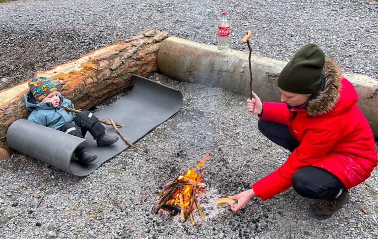 Grillarr korv i skog Barn sitter på en matta bredvid en eldstad medan en vuxen grillar vid elden.