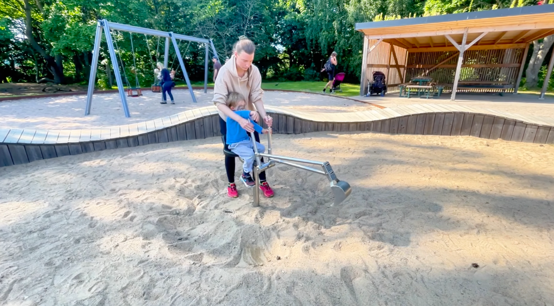 Vallarnas Friluftsområde Lekplatsen Kvinna som arbetar med att jämna ut sanden på en lekplats. Barn leker i bakgrunden.
