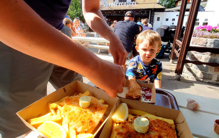 En hand sträcker sig efter en tallrik med mat medan ett barn sitter i bakgrunden.