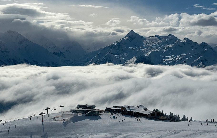 Fjällandskap med snö och moln som täcker dalarna, med majestätiska berg i bakgrunden.