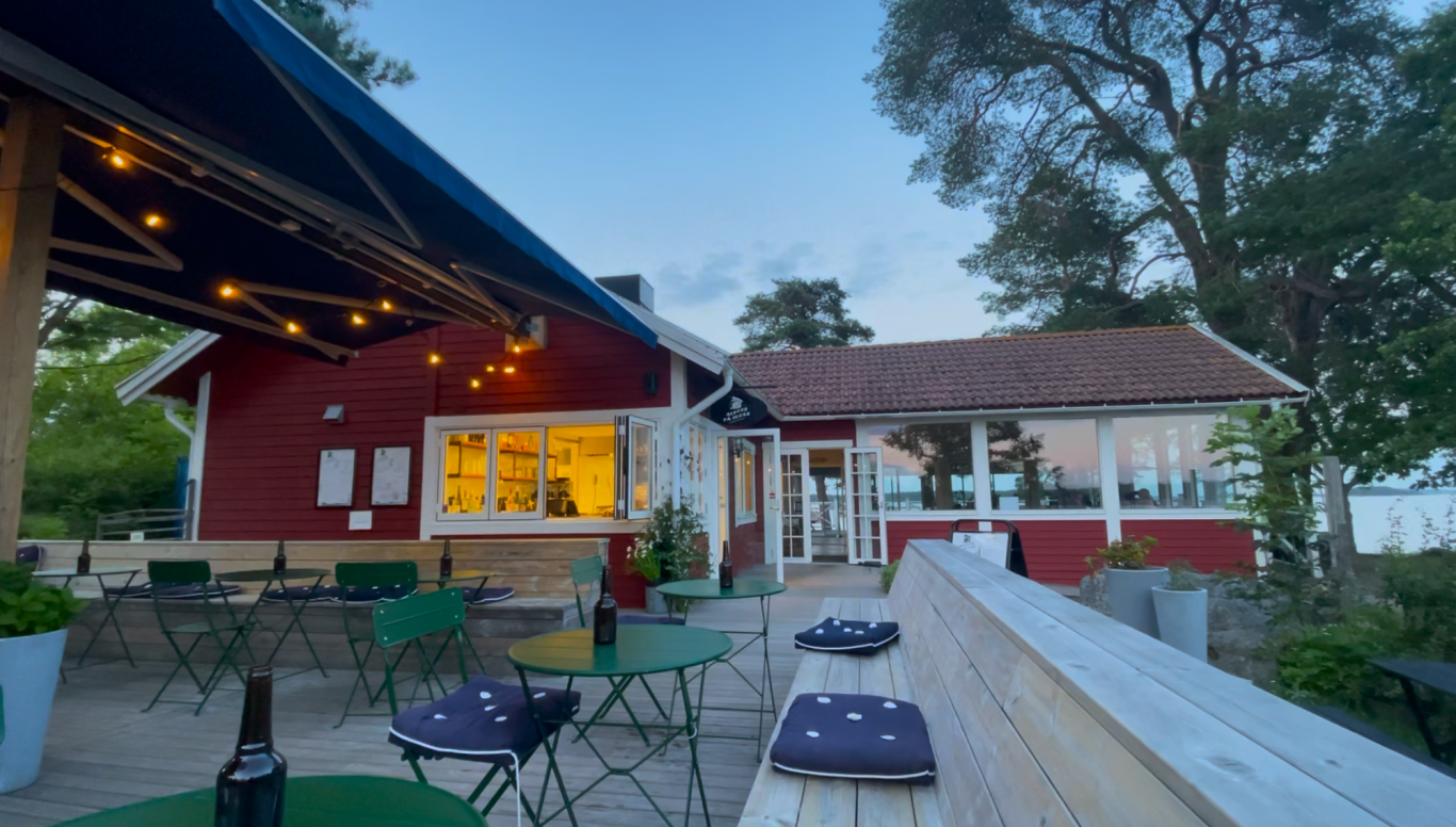 Algots på Udden Röd stuga med uteservering och belysning, omgiven av träd och grönska.