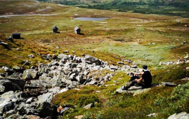 En person sitter på en klippa i ett öppet landskap med grön och brun vegetation.