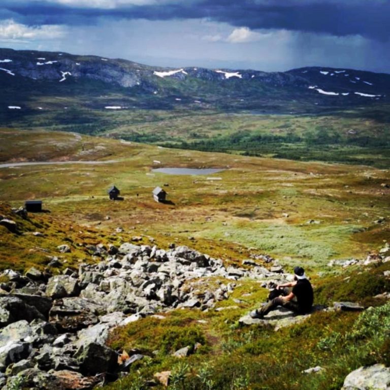 En person sitter på en sten i ett öppet fjällandskap med grön och gul vegetation.