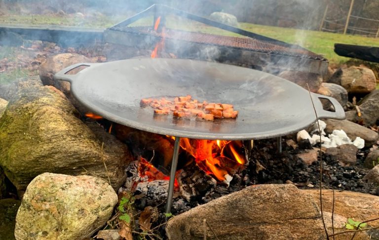 Grill med kött som tillagas över öppen eld, omgiven av stenar och rök.