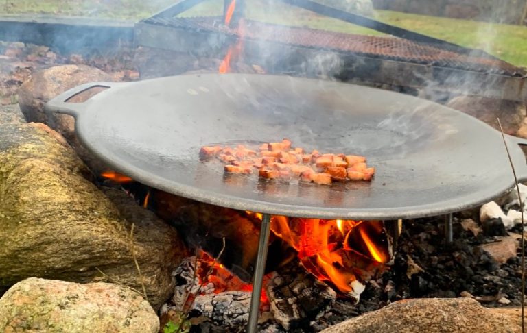 Kött som grillas på en stor platta över öppen eld med rök och stenar runtom.