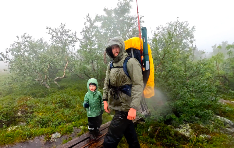 En vuxen och ett barn klädda i regnkläder, vandrar i dimmig skogsmiljö.