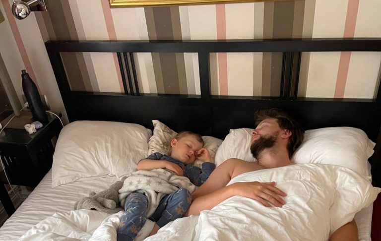 En man och ett barn sover tillsammans i en säng med vita lakan.