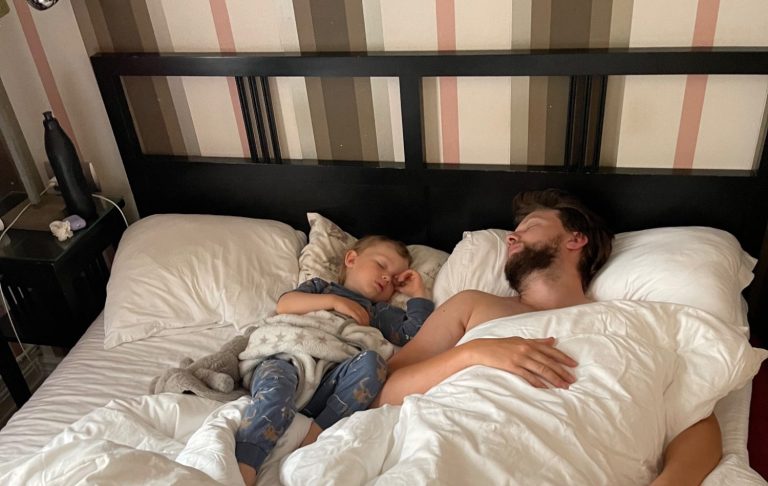 En vuxen och ett barn sover tillsammans i en säng med vit sängklädsel.