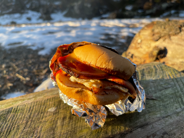 Falukorvsburgare med ägg, senap och ketchup på en bänk, omgiven av snö och natur.