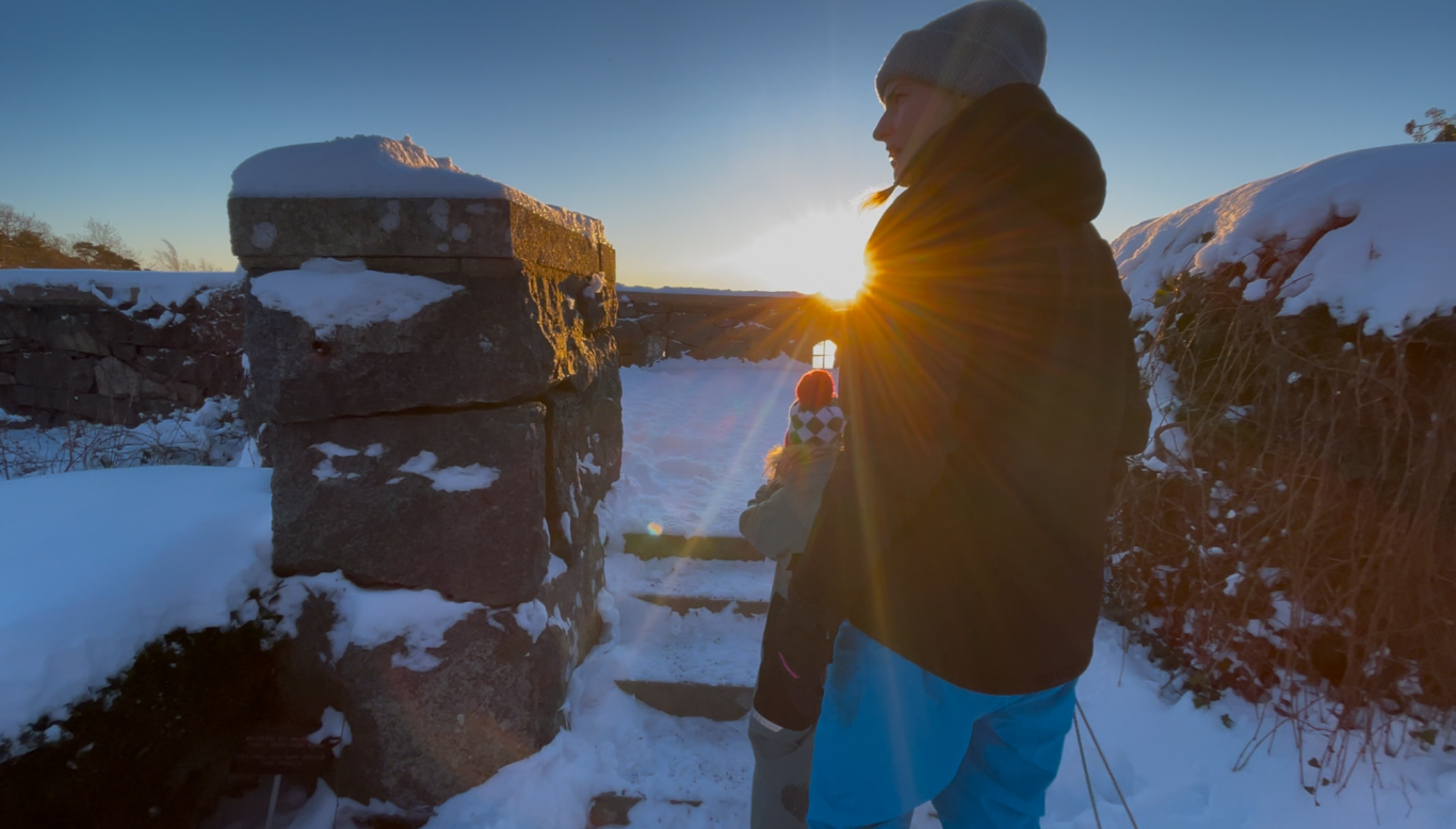 Solnedgång Person i vinterkläder står vid stenar, solnedgången lyser i bakgrunden.