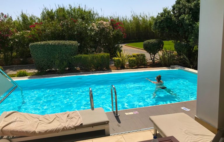 En person simmar i en klar blå pool, omgiven av grönska och blommor.