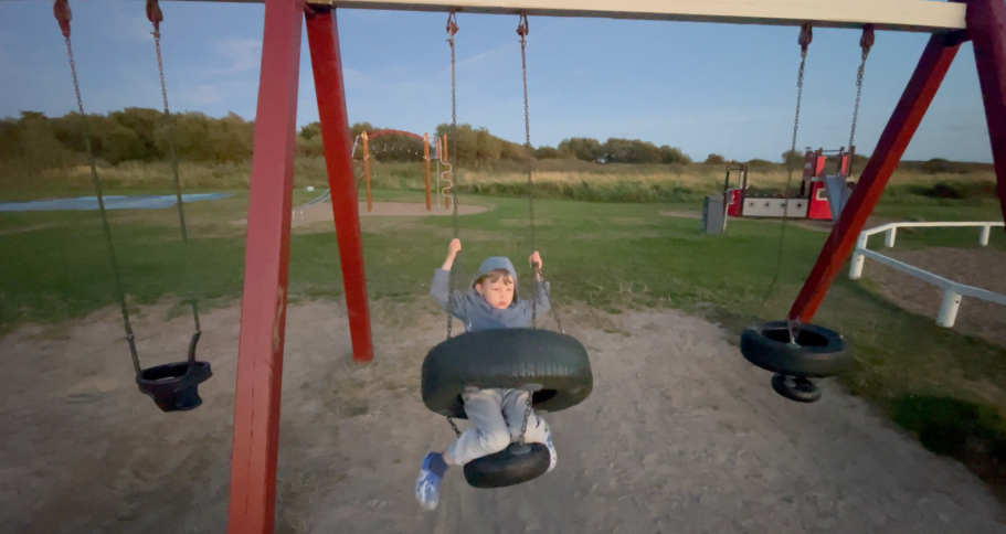 Barn leker på gungor i en park med grön gräsyta och träd i bakgrunden.