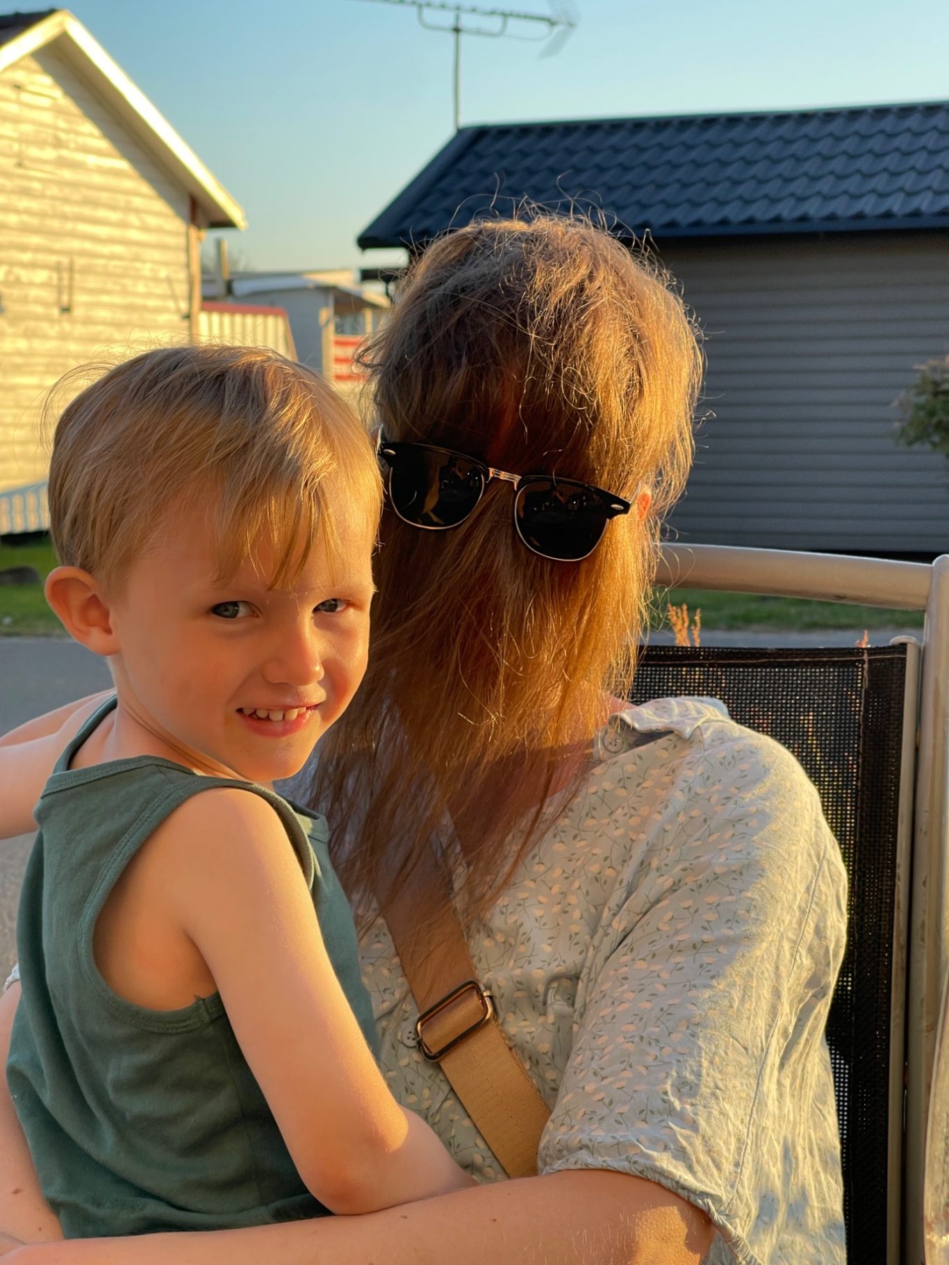 Ett Monster Barn sitter på en vuxens knä, båda ler mot kameran i solnedgång.