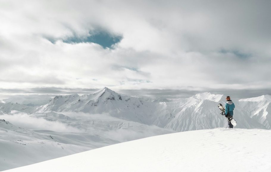 En person står på en snöklädd alptopp med fjärran berg i bakgrunden.
