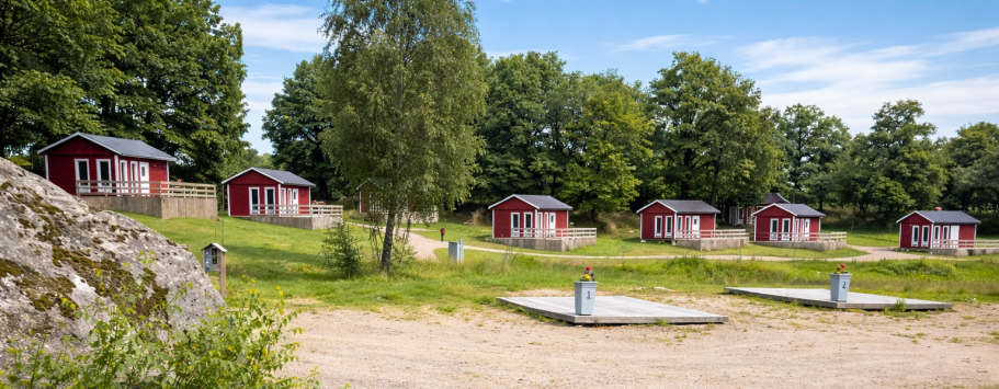 Röda stugor omgivna av grönska och träd, med en sandig mark och bryggor i förgrunden.