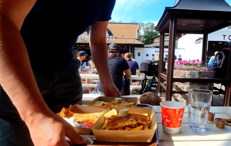 En person bär två matlådor på en utomhuscafé. Det är soligt och folk i bakgrunden.