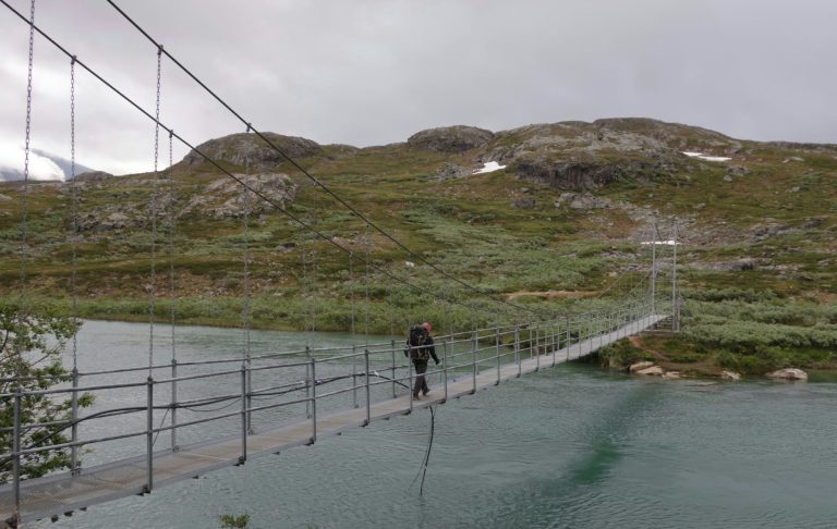 En Outdoorfestival Person går över en hängbro över en sjö omgiven av fjäll och grönska.