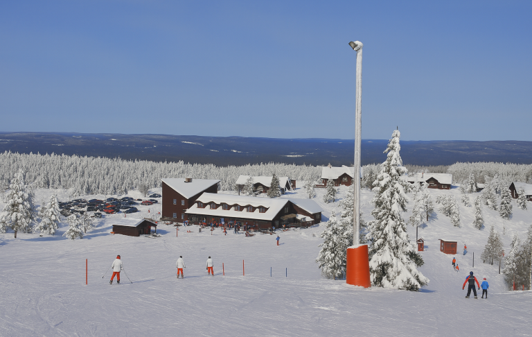 Snötäckt skidbacke med flera åkare och en stuga i bakgrunden. Klara himmelar.