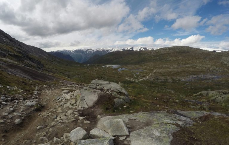 Bergigt landskap med stenig stig och vidsträckt dal under molnig himmel.