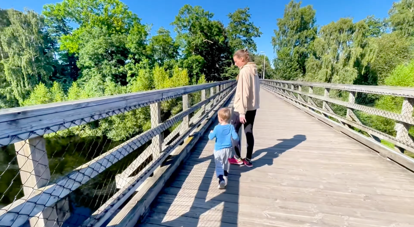 Bron till Vallarnas Friluftsområde En vuxen och ett barn går på en träbro omgiven av grönska.