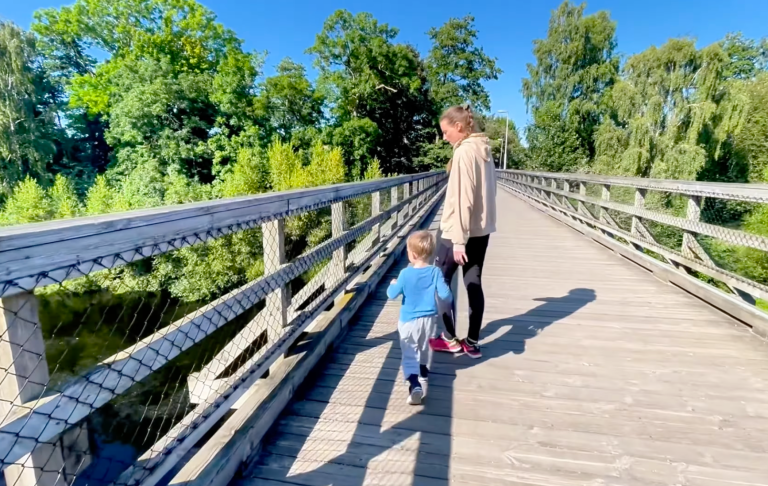 En vuxen och ett barn går hand i hand på en träbro omgiven av grönska.