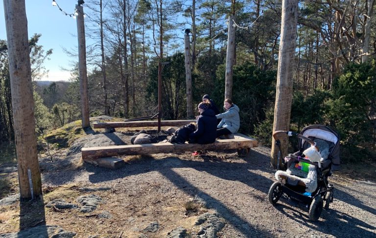 Två personer sitter vid en eldplats i skogen, en barnvagn står bredvid.