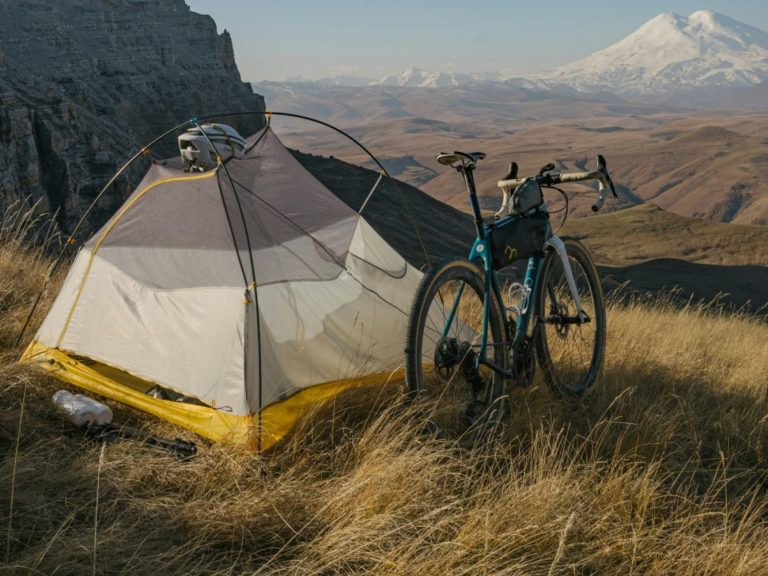 Tält på gräsbevuxen kulle med en cykel bredvid, fjärran berg i bakgrunden.