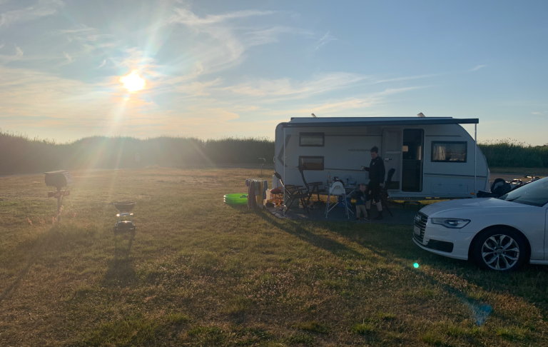 Campingplats med husvagn i solnedgång, stolar och en bil syns i förgrunden.