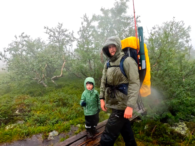 Vandrare med ryggsäck och litet barn i regnkläder i dimmig skogsmiljö.