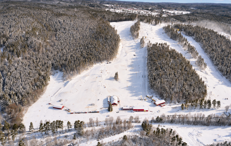 Snötäckt skidbacke med röda byggnader omgiven av täta granar.