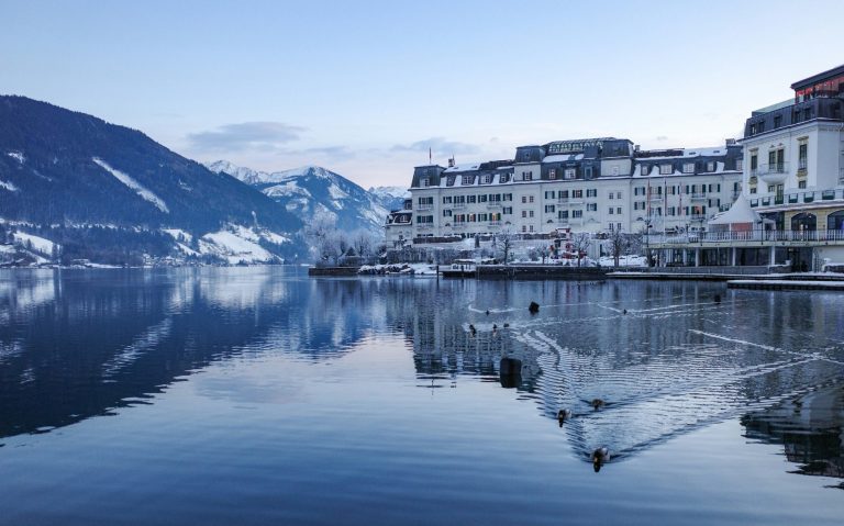 Lugnt vattenspegel med ett hotell och berg i bakgrunden, vid gryning.