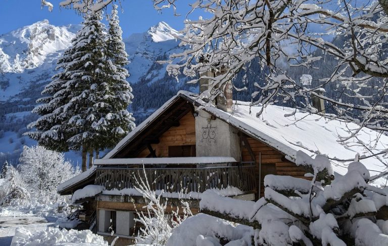 En stuga täckt av snö med stora alpberg och träd i bakgrunden.