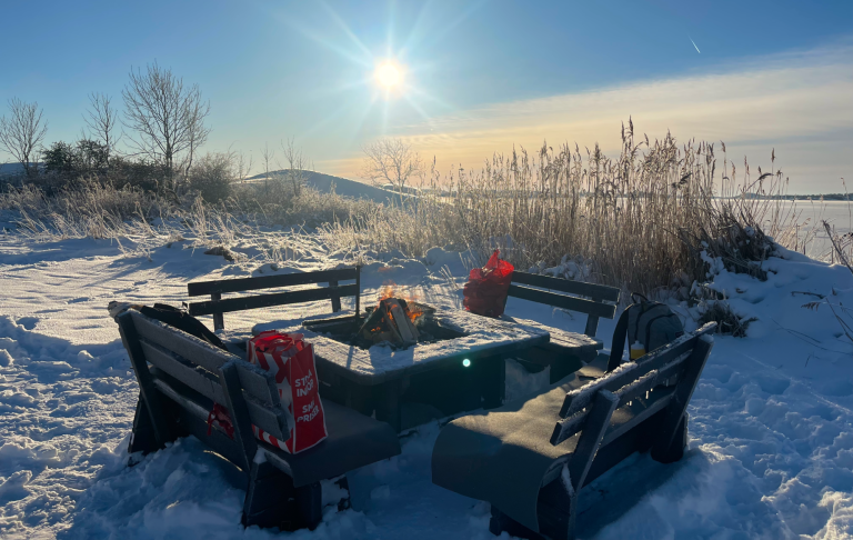 En snöig plats med bänkar och ett bord, solsken och vatten i bakgrunden.