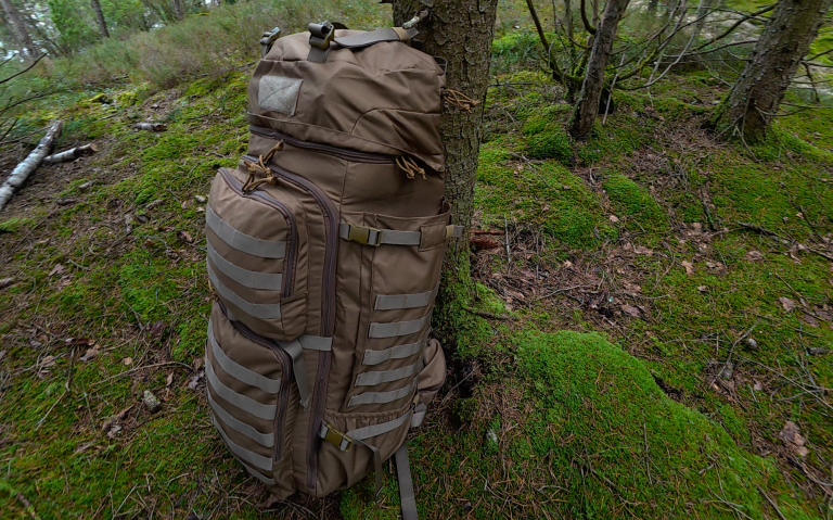 Brun ryggsäck lutad mot ett träd i en skog med mossa och skräp på marken.