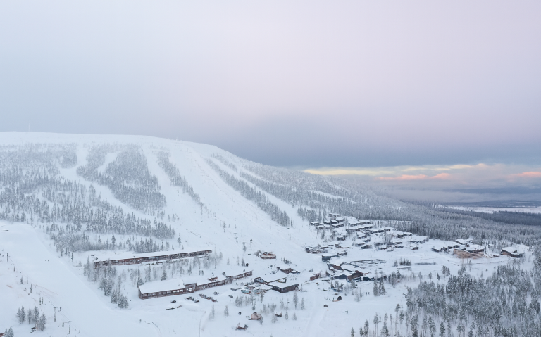 Snötäckt fjällandskap med stugor och skidbackar under en ljus himmel.