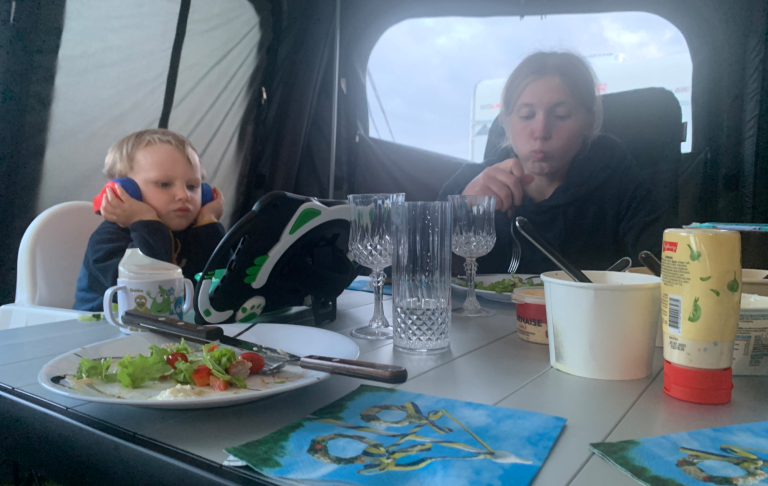 Barn sitter vid ett bord och äter, ser på en enhet i en campingmiljö.