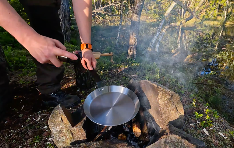 Person lagar mat över en öppen eld med rök som stiger i en skogsmiljö.