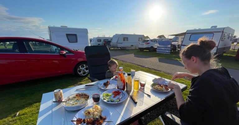 Familj äter middag vid campingplats under solnedgång. Campingar i bakgrunden.