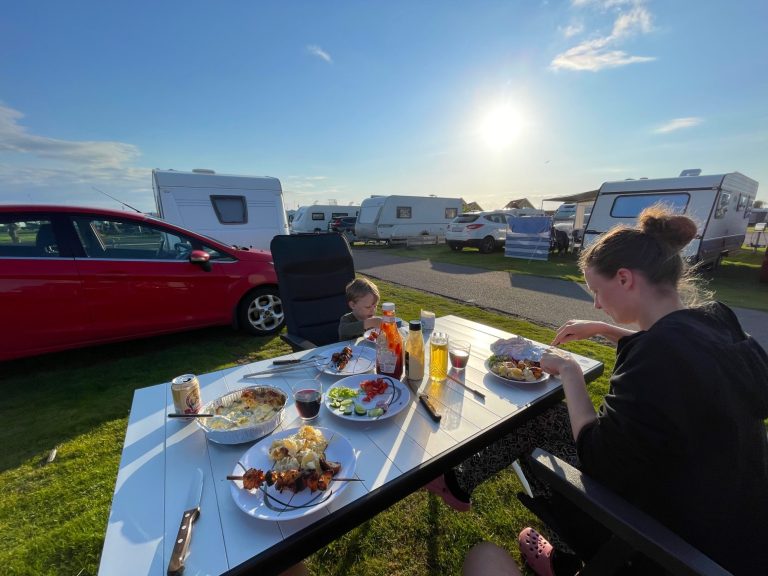 Familj äter middag vid campingplats under solnedgång. Campingar i bakgrunden.