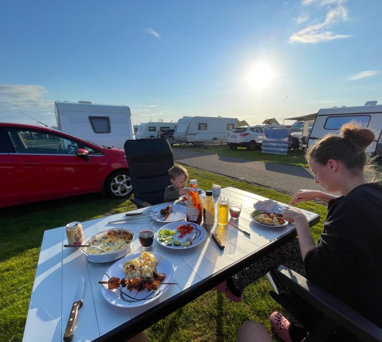Familj äter middag vid campingplats under solnedgång. Campingar i bakgrunden.