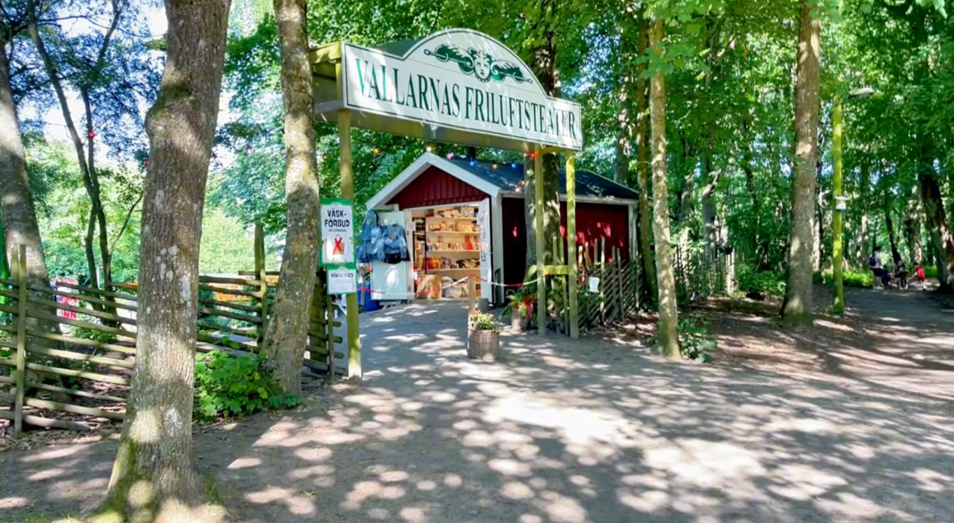 Entren till Vallarnas Friluftsteater Röd stuga med skylt i en grönskande skogsmiljö.