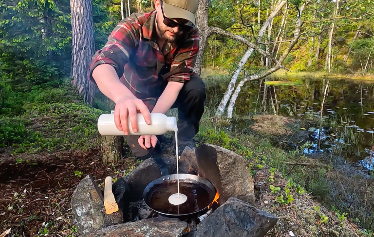 En person häller smet i en panna över öppen eld vid en sjö i skogen.