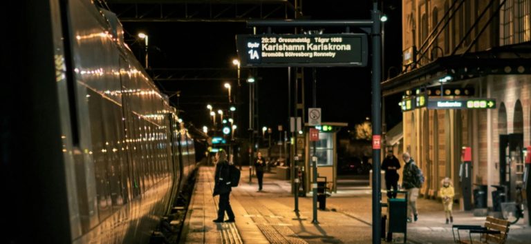 Mörk stadsscen med människor vid en tågstation och en informationsskylt.