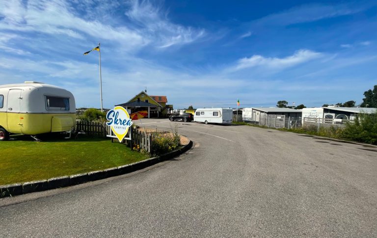 Väg mot en campingplats med husvagnar och grönska under klarblå himmel.