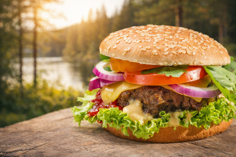 Saftig hamburgare med ost, grönsaker och sesambröd, framför en naturskön bakgrund.